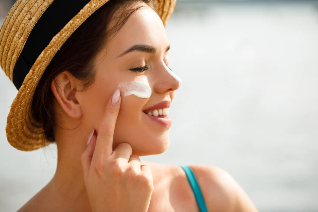 Woman in a sun hat applying skincare cream to her cheek outdoors
