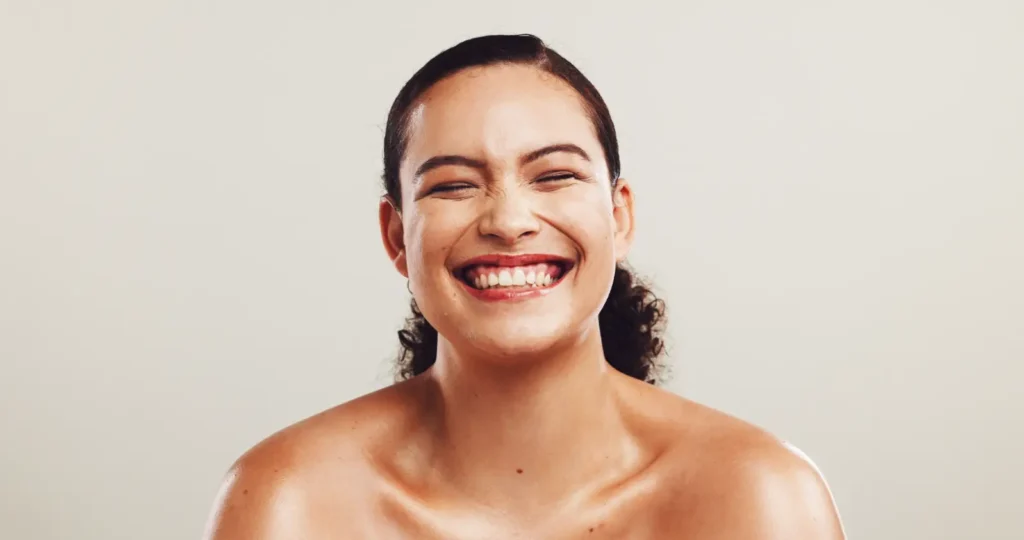 Smiling woman with bare shoulders and visible facial skin against a light neutral background.