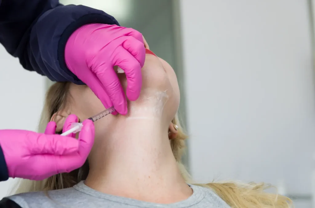 Practitioner in pink gloves administers an injection beneath a patient's chin.